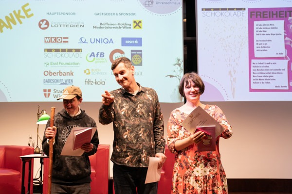 Drei Personen stehen auf einer Bühne und lächeln in die Kamera. Zwei Perosnen halten Urkunden in den Händen.